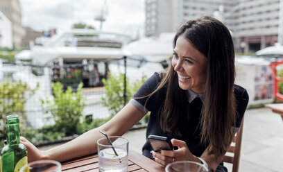 Glückliche junge Frau mit Smartphone und Getränk auf einer Terrasse - MGOF03892