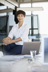 Portrait of confident businesswoman sitting on desk in office with laptop - RBF06859