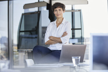 Portrait of confident businesswoman sitting on desk in office with laptop - RBF06860