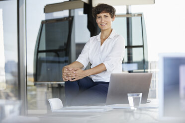 Portrait of confident businesswoman sitting on desk in office with laptop - RBF06861