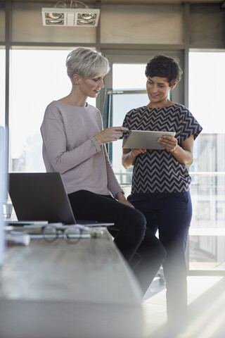 Zwei lächelnde Geschäftsfrauen teilen sich ein Tablet im Büro, lizenzfreies Stockfoto