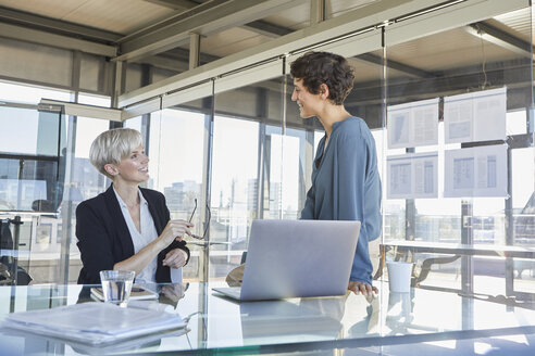 Zwei lächelnde Geschäftsfrauen, die am Schreibtisch im Büro diskutieren - RBF06895