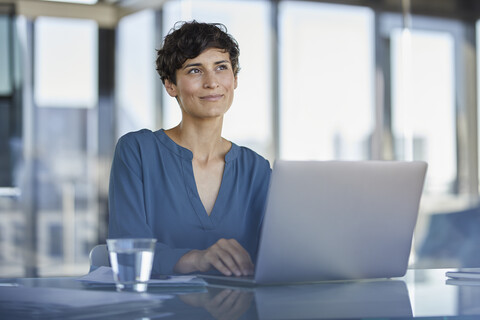 Lächelnde Geschäftsfrau sitzt am Schreibtisch im Büro mit Laptop, lizenzfreies Stockfoto