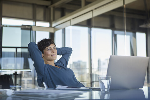 Lächelnde Geschäftsfrau sitzt am Schreibtisch im Büro mit Laptop und macht eine Pause, lizenzfreies Stockfoto