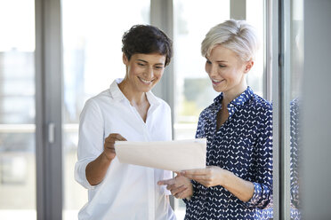 Two smiling businesswomen looking at document at the window in office - RBF06950