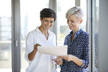 Zwei lächelnde Geschäftsfrauen betrachten ein Dokument am Fenster im Büro - RBF06950