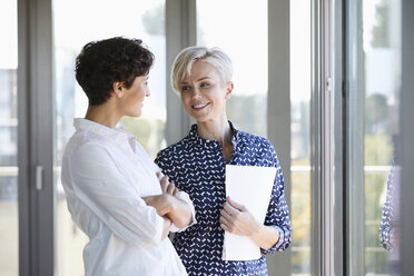 Zwei lächelnde Geschäftsfrauen im Gespräch am Fenster im Büro - RBF06953