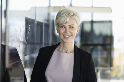 Portrait of smiling businesswoman next to chart on glass pane in office stock photo