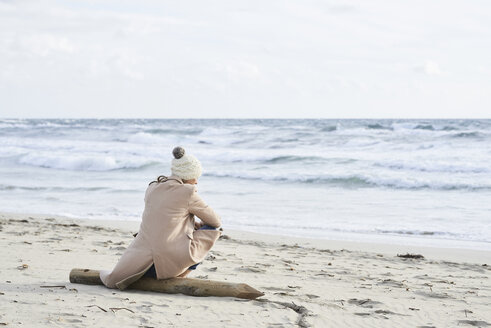 Spain, Menorca, back view of senior woman wearing bobble hat and coat sitting on the beach in winter - IGGF00695