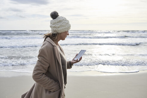 Spanien, Menorca, ältere Frau, die im Winter am Strand spazieren geht und ein E-Book liest - IGGF00702