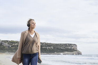 Spain, Menorca, senior woman using smartphone and wireless headphones on the beach in winter - IGGF00710