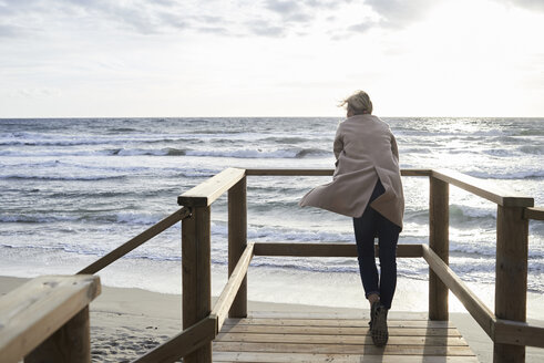 Spain, Menorca, back view of senior woman standing on boardwalk in winter looking at the sea - IGGF00715