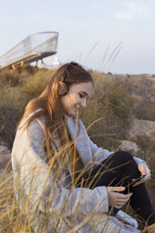 Spanien, Alicante, Santa Pola, Kap Santa Pola, junge Frau sitzt bei Sonnenuntergang auf einem Felsen und hört Musik mit ihrem Handy - GRSF00039
