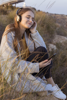 Spanien, Alicante, Santa Pola, Kap Santa Pola, junge Frau sitzt bei Sonnenuntergang auf einem Felsen und hört Musik mit ihrem Handy - GRSF00040