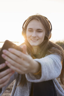 Glückliche junge Frau, die ein Selfie macht, während sie bei Sonnenuntergang Musik hört - GRSF00041