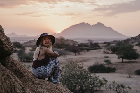 Namibia, Spitzkoppe, lachende Frau bei Sonnenuntergang auf einem Felsen sitzend, lizenzfreies Stockfoto