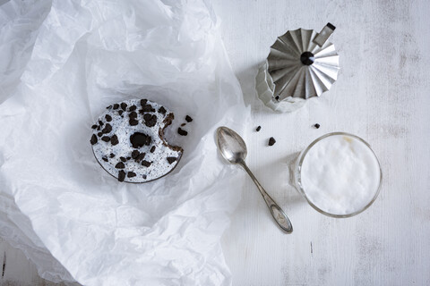 Gebissener Krapfen mit weißer Glasur und dunklen Schokoladensplittern, Mokkakanne, Teelöffel und Glas Cappuccino, lizenzfreies Stockfoto