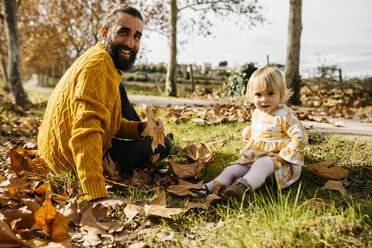 Father and daughter sitting on meadow with autumn leaves, morning day in the park - JRFF02259