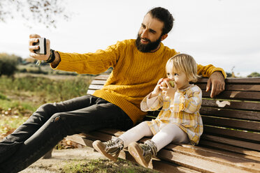 Father and daughter sitting on a bench in the park in autumn, father taking a selfie, daughter eating an apple - JRFF02273