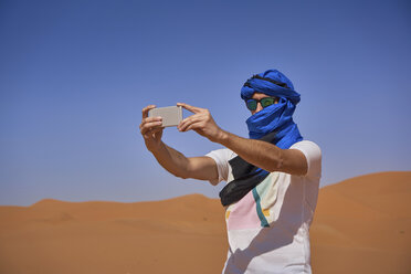 Morocco, man wearing sunglasses and blue tuban taking photo with smartphone in the desert - EPF00515