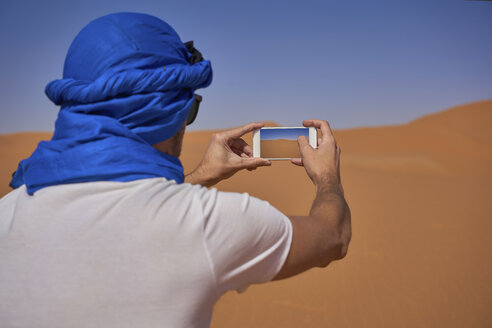 Morocco, back view of man wearing blue tuban taking photo with smartphone in the desert - EPF00516