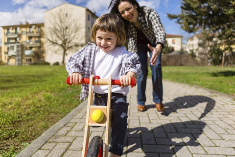 Mutter hinter glücklichem Kleinkind Sohn mit Balance Fahrrad auf einem Weg, lizenzfreies Stockfoto