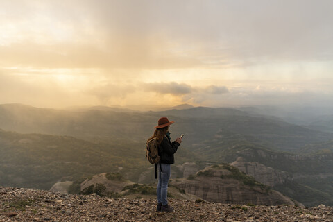Frau mit Rucksack, auf einem Berg stehend, mit Blick auf die Aussicht, mit Smartphone, lizenzfreies Stockfoto