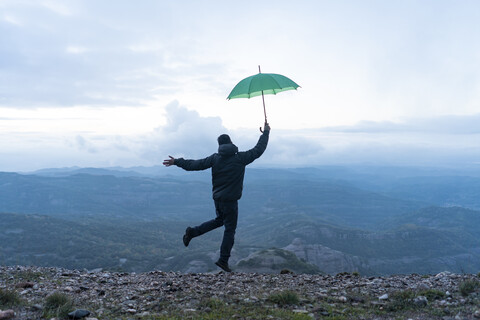 Mann, der vor Freude auf einem Berg springt und einen grünen Regenschirm hält, lizenzfreies Stockfoto