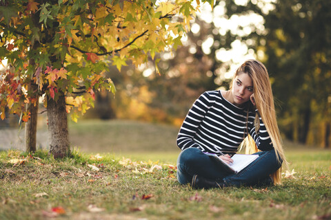Porträt einer nachdenklichen jungen Frau mit Notizbuch, die auf einer Wiese in einem Park im Herbst sitzt und sich Notizen macht, lizenzfreies Stockfoto