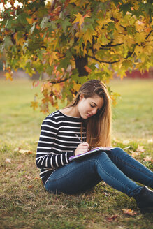 Junge Frau mit Notebook sitzt auf einer Wiese in einem Park im Herbst und macht sich Notizen - LOTF00007