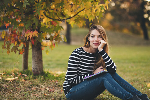 Young woman with notebook sitting on meadow in a park in autumn watching something - LOTF00008