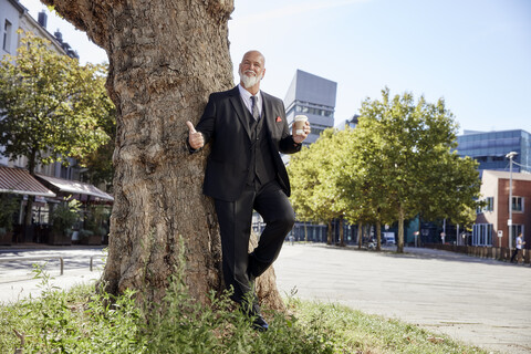 Eleganter Geschäftsmann lehnt an einem Baum in der Stadt und trinkt Kaffee, lizenzfreies Stockfoto