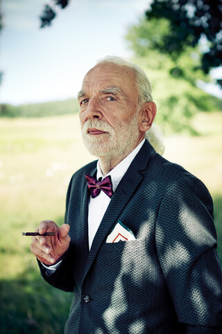 Porträt eines älteren Mannes mit Anzug, Fliege und Einstecktuch in der Natur, der Zigarillo raucht, lizenzfreies Stockfoto