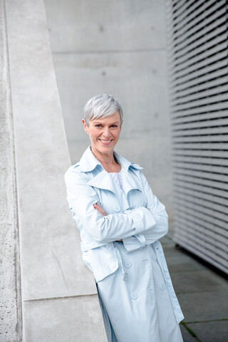 Porträt einer zufriedenen reifen Geschäftsfrau, die sich an eine Wand lehnt und einen hellblauen Trenchcoat trägt, lizenzfreies Stockfoto