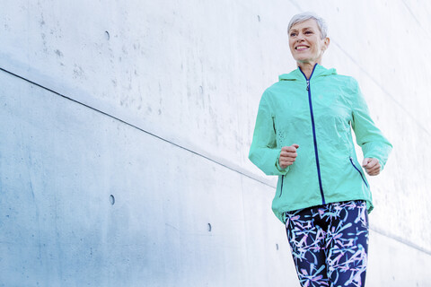 Smiling mature woman jogging along concrete wall stock photo