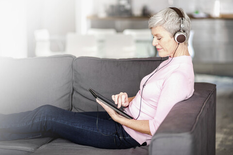 Mature woman sitting on couch using digital tablet while listening music with headphones stock photo