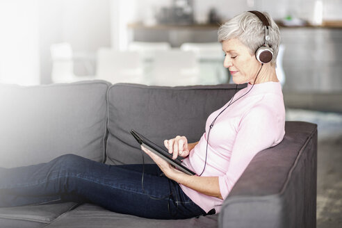 Ältere Frau sitzt auf der Couch und benutzt ein digitales Tablet, während sie mit Kopfhörern Musik hört - VWF00041