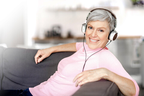 Porträt einer entspannten reifen Frau, die auf der Couch sitzt und mit Kopfhörern Musik hört, lizenzfreies Stockfoto