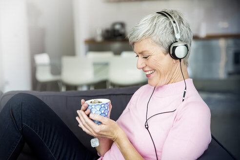 Lachende reife Frau sitzt mit einer Tasse Tee auf der Couch und hört Musik mit Kopfhörern - VWF00045