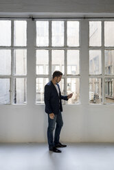 Businessman standing in a loft at the window using cell phone - MAUF02175