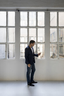 Geschäftsmann steht in einem Loft am Fenster und benutzt ein Mobiltelefon - MAUF02175