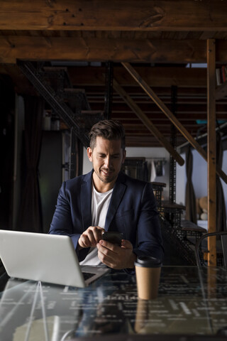 Lächelnder Geschäftsmann mit Mobiltelefon und Laptop in einem Studio-Loft, lizenzfreies Stockfoto