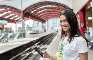 Großbritannien, London, Porträt einer lächelnden jungen Frau mit Handy in der Hand in einem Bahnhof - MGOF03915