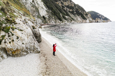 Italy, Elba, woman with red coat taking a photo at beach, aerial view with drone - FBAF00222