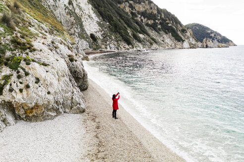 Italien, Elba, Frau mit rotem Mantel beim Fotografieren am Strand, Luftaufnahme mit Drohne - FBAF00222