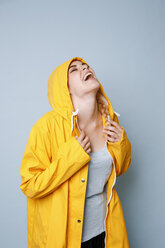 Laughing young woman wearing yellow rain coat in front of blue background - GRSF00048