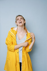 Portrait of laughing young woman wearing yellow rain coat in front of blue background - GRSF00049