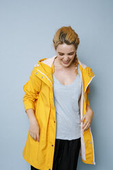 Lachende junge Frau mit gelbem Regenmantel vor blauem Hintergrund - GRSF00050