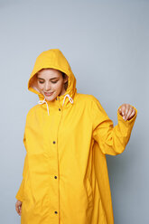 Smiling young woman wearing yellow rain coat in front of blue background dancing - GRSF00052