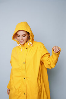 Lächelnde junge Frau in gelbem Regenmantel vor blauem Hintergrund tanzend - GRSF00052